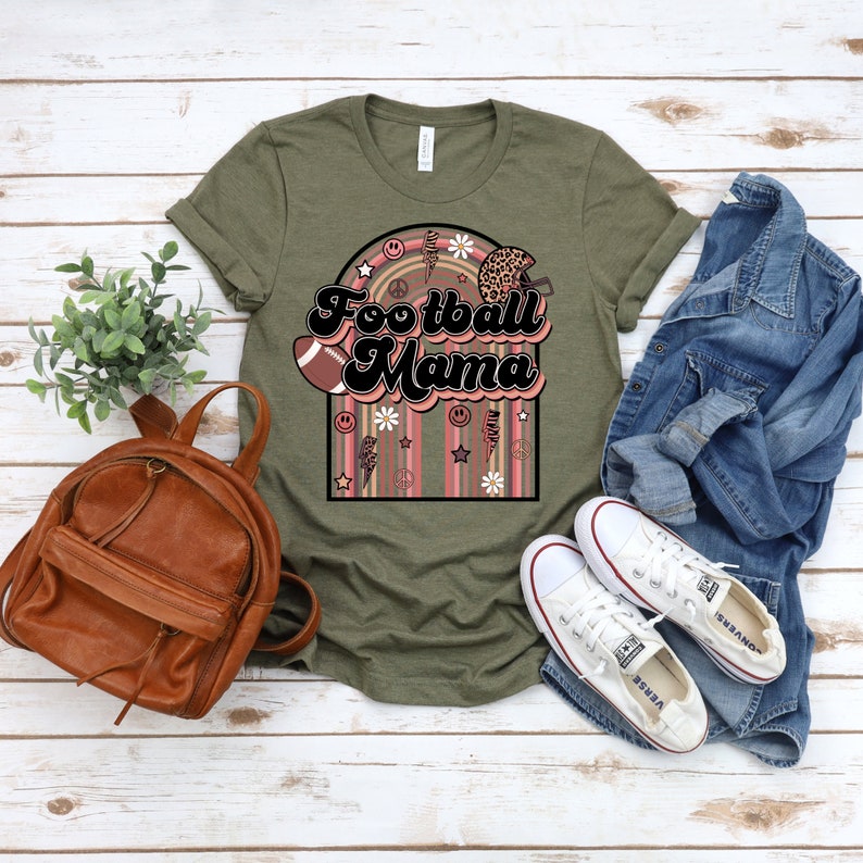 May include: A green t-shirt featuring the text "Football Mama" in a retro font. The design includes a football, daisies, peace signs, and lightning bolts.