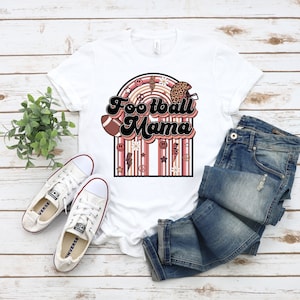 May include: White t-shirt featuring a retro-style graphic that reads "Football Mama" in brown and black lettering. The graphic includes a football, a helmet, daisies, peace signs, and lightning bolts. The graphic is surrounded by pink and brown stripes.