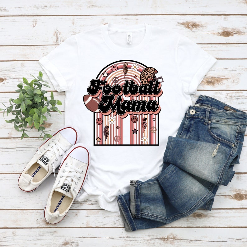 May include: White t-shirt featuring a retro-style graphic that reads "Football Mama" in brown and black lettering. The graphic includes a football, a helmet, daisies, peace signs, and lightning bolts. The graphic is surrounded by pink and brown stripes.