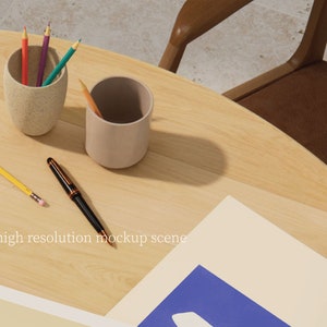 May include: A light brown wooden table with two beige pencil holders, one containing coloured pencils and the other empty. A yellow pencil and a black pen are on the table. A white sheet of paper with a blue rectangle on top is on the table. The text "high resolution mockup scene" is printed on the paper.