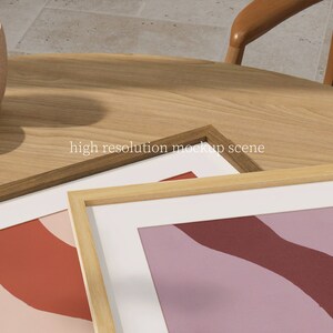 May include: Two wooden picture frames with white mats and abstract art prints in shades of pink, red, and purple. The frames are on a wooden table.