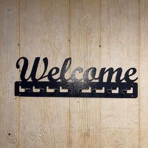 Puede incluir: Un letrero de bienvenida de metal negro con ganchos para colgar objetos. El letrero está montado en una pared de madera.