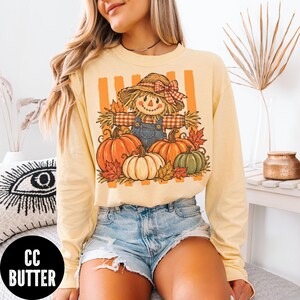 May include: A pale yellow long-sleeve shirt featuring a graphic print of a scarecrow wearing a straw hat with a red plaid bow, standing behind three pumpkins in orange, white, and green, set against a background of vertical orange stripes.