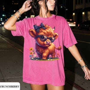 Puede incluir: Camiseta rosa con una vaca de las Tierras Altas de dibujos animados con gafas de sol y un lazo a cuadros. La vaca est&aacute; rodeada de flores de colores. El texto "CRUNCHBERRY" est&aacute; en la parte inferior.