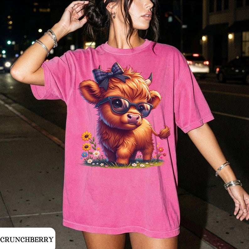 Puede incluir: Camiseta rosa con una vaca de las Tierras Altas de dibujos animados con gafas de sol y un lazo a cuadros. La vaca est&aacute; rodeada de flores de colores. El texto "CRUNCHBERRY" est&aacute; en la parte inferior.