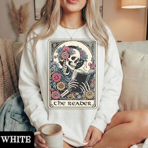 May include: A white long-sleeve sweatshirt featuring a graphic print of a skeleton reading a book, surrounded by colorful flowers. The design is framed in a rectangular border with the text THE READER printed in black at the bottom of the graphic.
