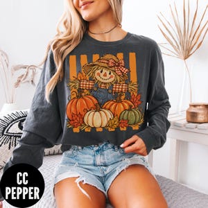 May include: A charcoal gray long-sleeve sweatshirt featuring a graphic print of a scarecrow wearing a straw hat with a red plaid bow, dressed in blue overalls, surrounded by orange and green pumpkins and autumn leaves against orange vertical stripes.