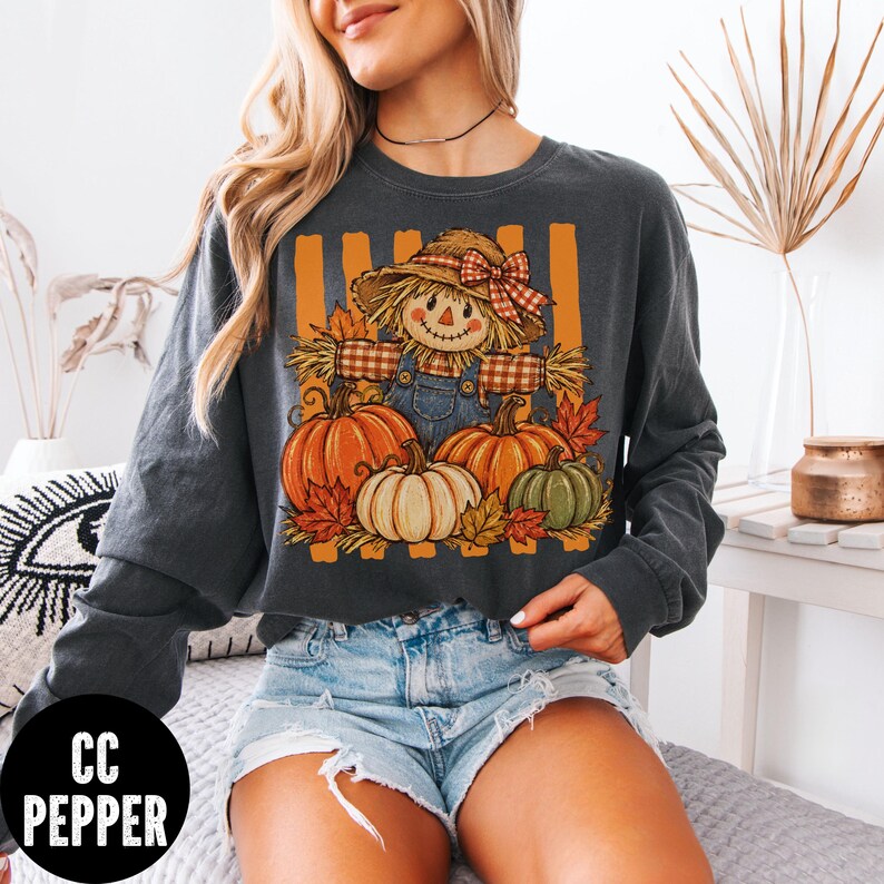May include: A charcoal gray long-sleeve sweatshirt featuring a graphic print of a scarecrow wearing a straw hat with a red plaid bow, dressed in blue overalls, surrounded by orange and green pumpkins and autumn leaves against orange vertical stripes.