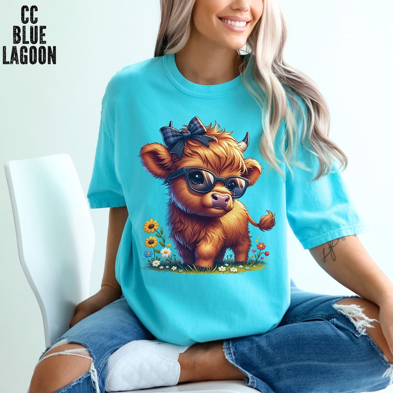 Puede incluir: Camiseta azul claro con una ilustraci&oacute;n de dibujos animados de una vaca de las Tierras Altas con gafas de sol y un lazo. La vaca es marr&oacute;n con flores amarillas y hierba verde. El texto "CC BLUE LAGOON" est&aacute; en la esquina superior izquierda.