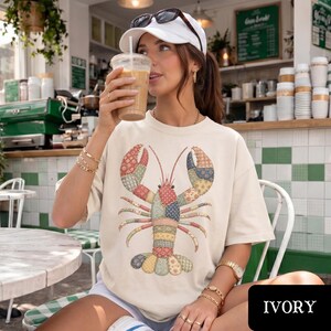 May include: An ivory short-sleeve t-shirt featuring a large, central graphic of a lobster composed of various colorful, patterned quilt-like patches. The design includes floral, polka dot, and geometric prints in shades of blue, pink, and green.