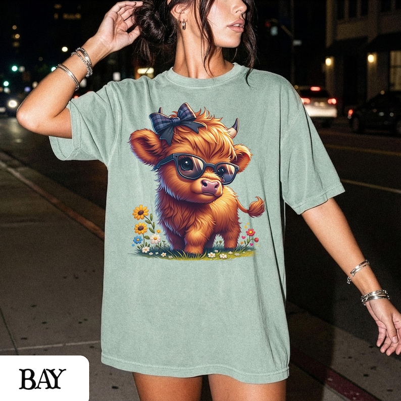 Puede incluir: Camiseta verde claro con una ilustraci&oacute;n de dibujos animados de una vaca de las Tierras Altas con gafas de sol y un lazo. La vaca es marr&oacute;n con la nariz rosa y est&aacute; rodeada de flores. La palabra "BAY" est&aacute; abajo.