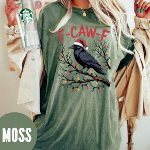 May include: Moss green t-shirt with a graphic of a black crow wearing a Santa hat perched on a branch decorated with Christmas lights. The text "F-CAW-F" is above the crow.