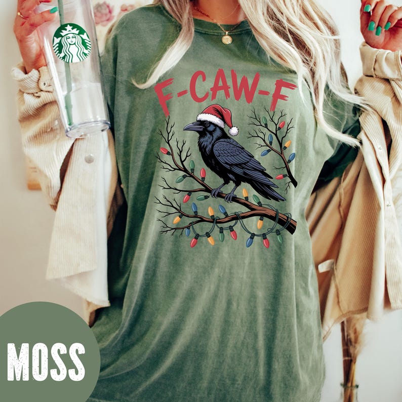 May include: Moss green t-shirt with a graphic of a black crow wearing a Santa hat perched on a branch decorated with Christmas lights. The text "F-CAW-F" is above the crow.