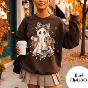 May include: A dark brown sweatshirt featuring a whimsical ghost design with a bow, holding a mug and gingerbread man. The ghost is adorned with festive patterns and string lights. The words "Dark Chololate" are written in a script font on the bottom right.