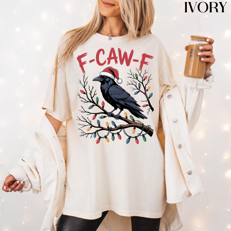 May include: Ivory-colored t-shirt featuring a black crow wearing a Santa hat, perched on a branch adorned with colorful Christmas lights. The text "F-CAW-F" is printed above the crow in red. The shirt is a relaxed fit.
