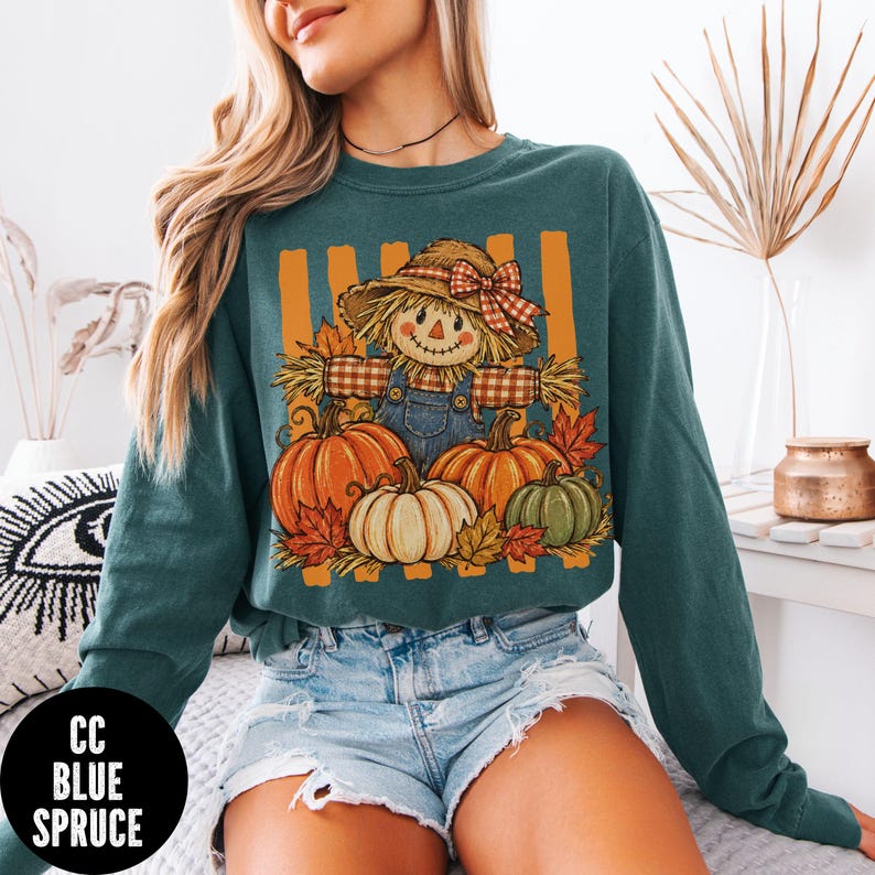 May include: A dark teal long-sleeve crewneck sweatshirt featuring a graphic print of a scarecrow wearing a straw hat with a red plaid bow, surrounded by orange, white, and green pumpkins and autumn leaves against vertical orange stripes.
