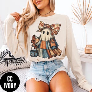 May include: Cream-colored long-sleeve sweatshirt featuring a whimsical ghost design. The ghost wears a patchwork outfit with a floral bow and carries a blue jack-o'-lantern bucket. The sweatshirt has the text "CC IVORY" in a black circle.