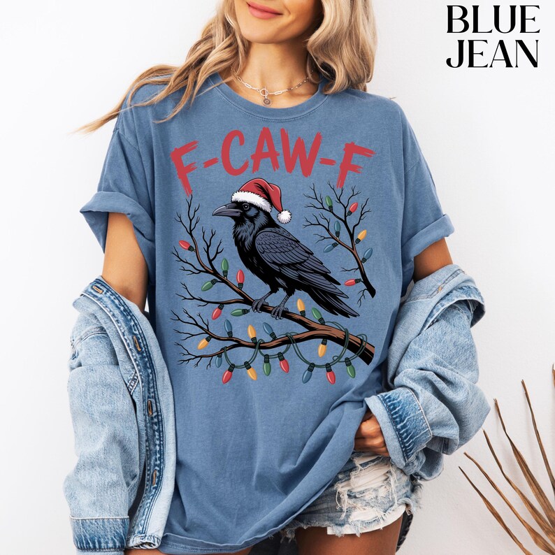May include: Blue t-shirt with a graphic of a black crow wearing a Santa hat perched on a branch decorated with Christmas lights. The text "F-CAW-F" is printed above the crow in red.