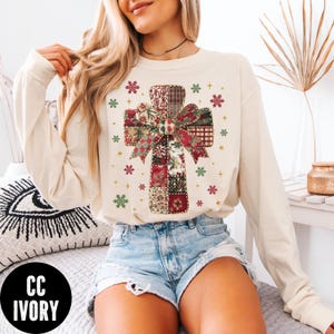 May include: Cream-colored long-sleeve sweatshirt featuring a patchwork cross design with a bow. The cross is made of various patterned fabrics in red, green, and cream colors. The design is surrounded by small snowflakes. The text "CC IVORY" is in a black circle.