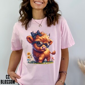 Puede incluir: Camiseta rosa claro con una ilustraci&oacute;n de dibujos animados de una vaca de las Tierras Altas con gafas de sol y un lazo a cuadros. La vaca est&aacute; rodeada de flores coloridas. El texto "CC BLOSSOM" est&aacute; impreso en negro en la parte inferior izquierda.