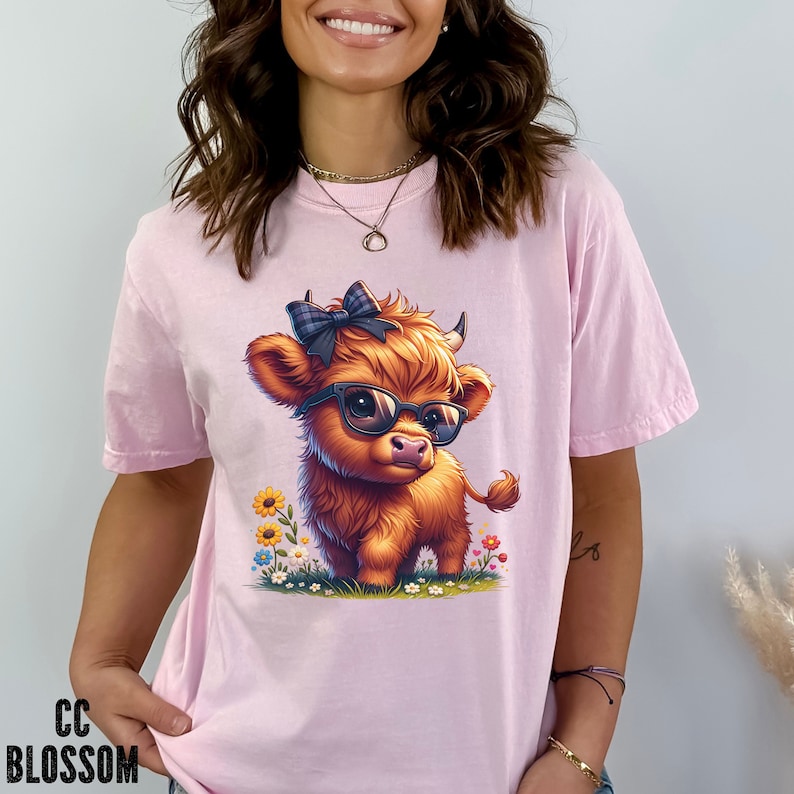 Puede incluir: Camiseta rosa claro con una ilustraci&oacute;n de dibujos animados de una vaca de las Tierras Altas con gafas de sol y un lazo a cuadros. La vaca est&aacute; rodeada de flores coloridas. El texto "CC BLOSSOM" est&aacute; impreso en negro en la parte inferior izquierda.