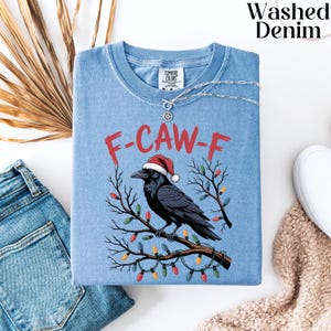 May include: Light blue t-shirt with a graphic of a black crow wearing a Santa hat, perched on a branch decorated with Christmas lights. The text "F-CAW-F" is printed above the crow in red. The shirt is folded with a silver necklace.