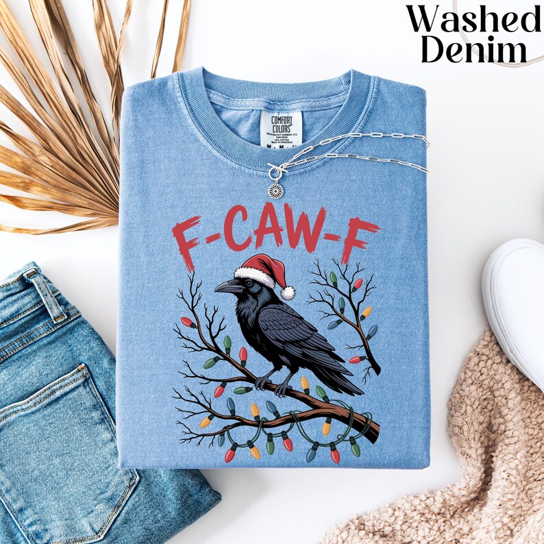 May include: Light blue t-shirt with a graphic of a black crow wearing a Santa hat, perched on a branch decorated with Christmas lights. The text "F-CAW-F" is printed above the crow in red. The shirt is folded with a silver necklace.