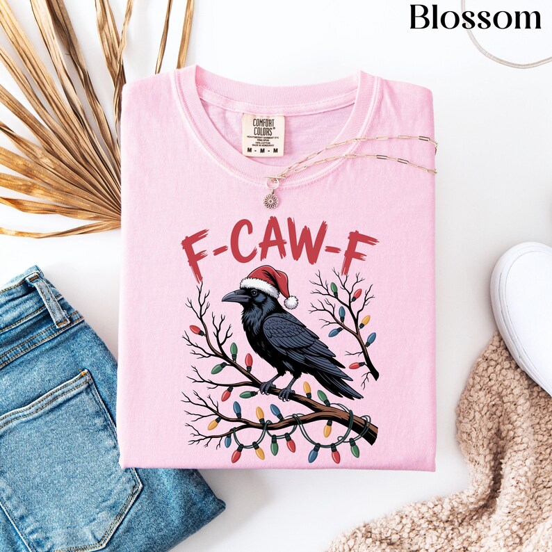 May include: A light pink t-shirt with a graphic of a black crow wearing a Santa hat perched on a branch decorated with Christmas lights. The text "F-CAW-F" is above the crow. A gold necklace and blue jeans are next to the shirt.