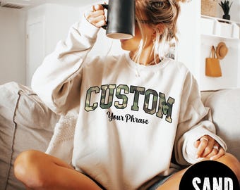 May include: A sand-colored sweatshirt with the word "CUSTOM" in a camouflage design and the phrase "Your Phrase" underneath. The sweatshirt is being worn by a person holding a black mug. A black circle with the word "SAND" is in the bottom right corner.