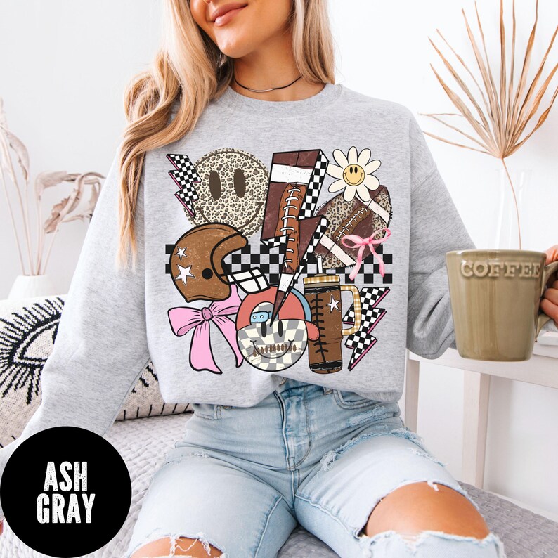 May include: Ash gray sweatshirt with a graphic design featuring footballs, a helmet, a daisy, and a smiley face. The design incorporates checkered patterns, lightning bolts, and a pink bow. The word "COFFEE" is printed on a mug.