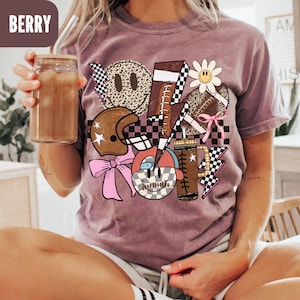 K&ouml;nnte beinhalten: Beerenfarbenes T-Shirt mit einem grafischen Design mit einem Football, Helm, Smiley-Gesichtern und Blitz. Das Design enth&auml;lt eine Mischung aus Mustern, darunter Leopardenmuster und Schachbrettflaggen. Ein Glas mit einem Getr&auml;nk wird in der Hand gehalten.