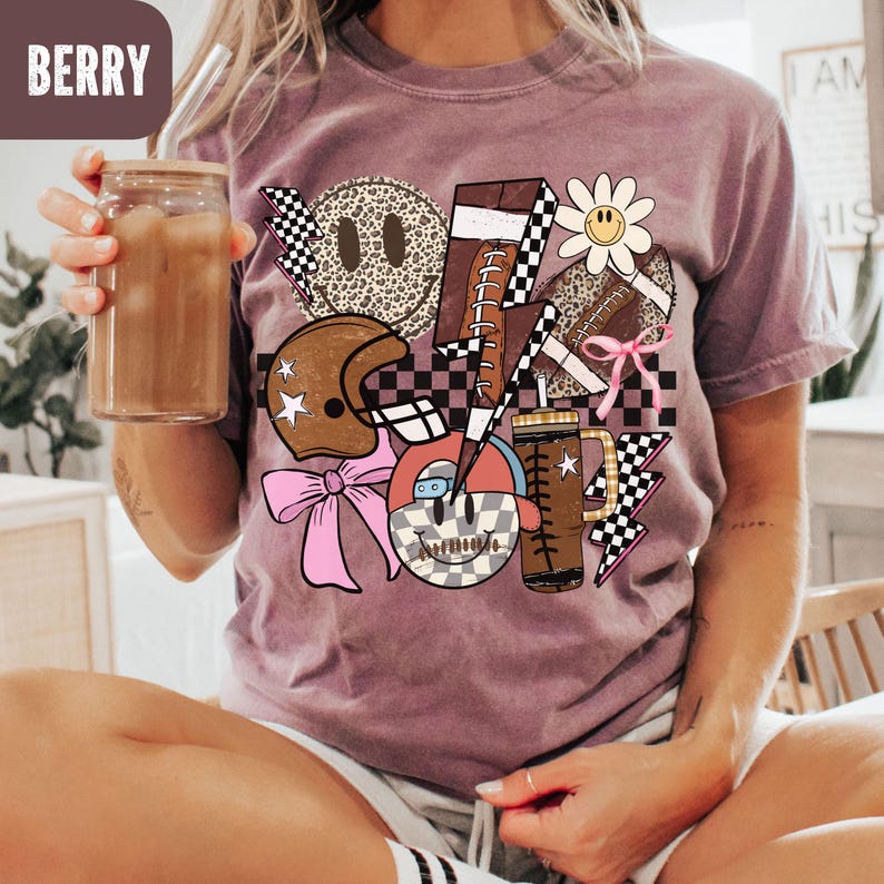 K&ouml;nnte beinhalten: Beerenfarbenes T-Shirt mit einem grafischen Design mit einem Football, Helm, Smiley-Gesichtern und Blitz. Das Design enth&auml;lt eine Mischung aus Mustern, darunter Leopardenmuster und Schachbrettflaggen. Ein Glas mit einem Getr&auml;nk wird in der Hand gehalten.