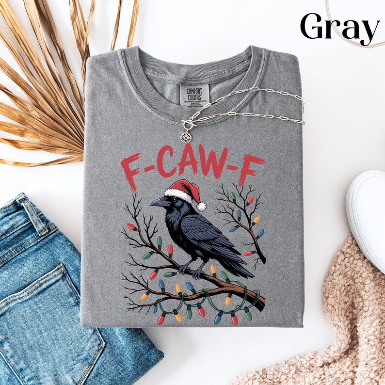 May include: Gray Comfort Colors t-shirt with a graphic of a black crow wearing a Santa hat, perched on a branch decorated with Christmas lights. The text "F-CAW-F" is above the crow. A silver necklace is draped across the shirt.