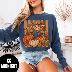 May include: A blue long-sleeve sweatshirt featuring a graphic print of a scarecrow wearing a straw hat with a red plaid bow, surrounded by orange and green pumpkins and autumn leaves against a background of vertical orange stripes.