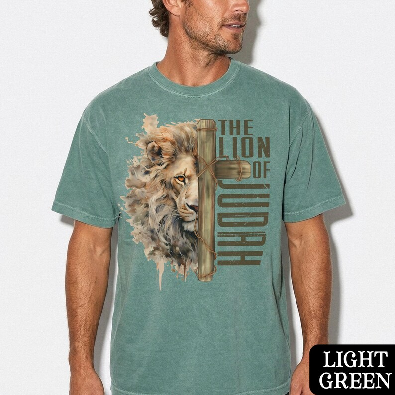 K&ouml;nnte beinhalten: Hellgr&uuml;nes T-Shirt mit einem L&ouml;wengesicht und einem Kreuz-Grafik mit dem Text "THE LION OF JUDAH" in Braun. Die L&ouml;wenm&auml;hne ist mit Braun- und Beiget&ouml;nen detailliert. Das Shirt hat eine einfarbige hellgr&uuml;ne Farbe.