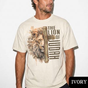 K&ouml;nnte beinhalten: Elfenbeinfarbenes T-Shirt mit einer Aquarellillustration eines L&ouml;wengesichts und eines Holzkreuzes. Der Text "THE LION OF JUDAH" ist vertikal auf der rechten Seite des Kreuzes aufgedruckt. Das T-Shirt hat kurze &Auml;rmel und einen Rundhalsausschnitt.