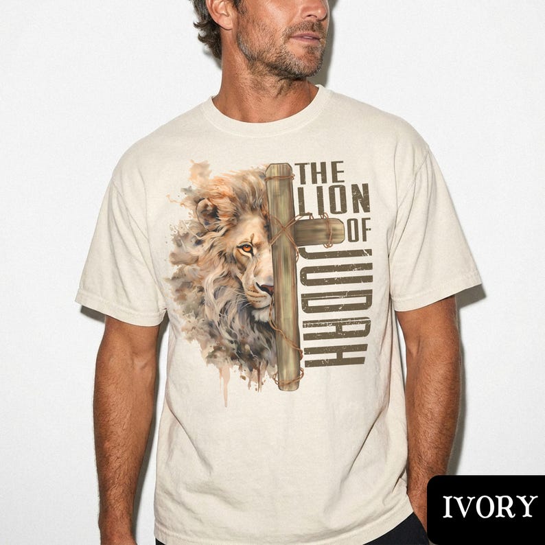 K&ouml;nnte beinhalten: Elfenbeinfarbenes T-Shirt mit einer Aquarellillustration eines L&ouml;wengesichts und eines Holzkreuzes. Der Text "THE LION OF JUDAH" ist vertikal auf der rechten Seite des Kreuzes aufgedruckt. Das T-Shirt hat kurze &Auml;rmel und einen Rundhalsausschnitt.