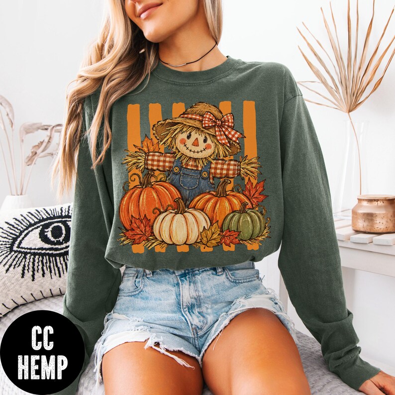 May include: A dark olive green long-sleeve sweatshirt featuring a graphic print of a scarecrow wearing a plaid shirt and a straw hat with a red checkered bow, surrounded by orange and green pumpkins and autumn leaves against vertical orange stripes.