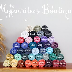 Puede incluir: Una pir&aacute;mide de camisetas enrolladas en varios colores, incluyendo negro, azul, verde y rosa. Cada camiseta est&aacute; etiquetada con el nombre de su color. El texto "Myfavritees Boutique" se muestra en una fuente cursiva en la parte superior.