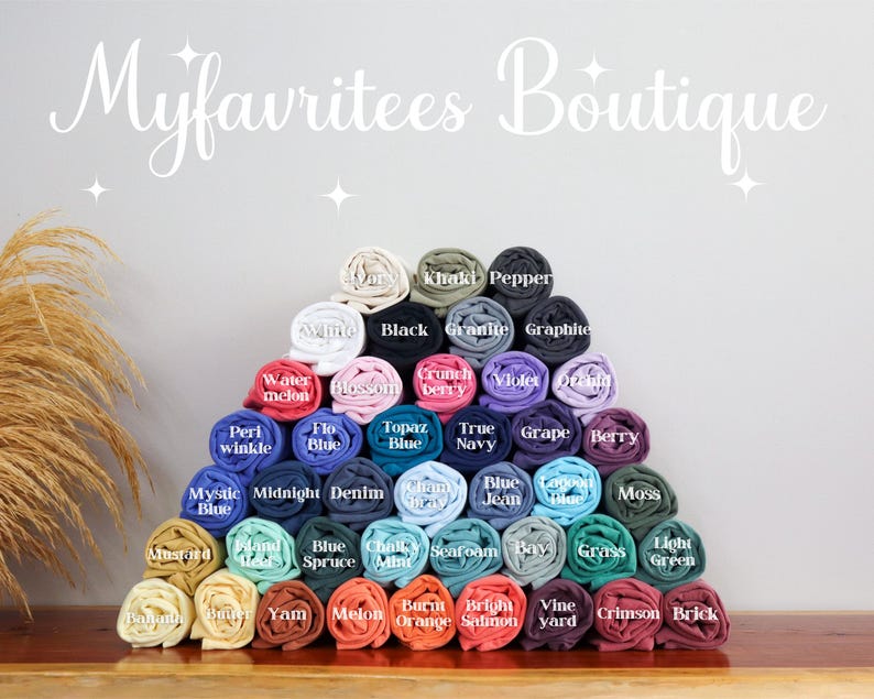 Puede incluir: Una pir&aacute;mide de camisetas enrolladas en varios colores, incluyendo negro, azul, verde y rosa. Cada camiseta est&aacute; etiquetada con el nombre de su color. El texto "Myfavritees Boutique" se muestra en una fuente cursiva en la parte superior.