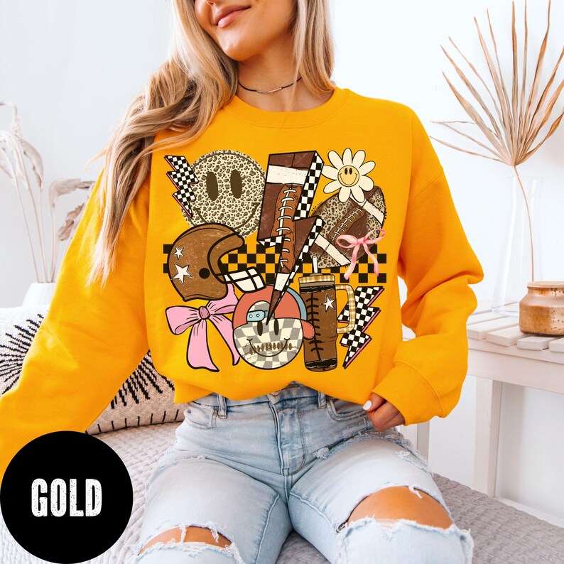 May include: Mustard yellow sweatshirt with a graphic design featuring footballs, lightning bolts, a football helmet, and a smiley face. The word "GOLD" is printed in a black circle at the bottom.
