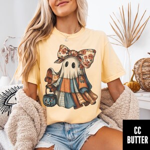 May include: Yellow t-shirt featuring a whimsical ghost design. The ghost wears a patchwork outfit and a floral bow, holding a jack-o'-lantern bucket. The design has a vintage, fall-themed aesthetic, perfect for Halloween or autumn.