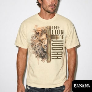 K&ouml;nnte beinhalten: Cremefarbenes T-Shirt mit einer Aquarellillustration eines L&ouml;wengesichts und eines Holzkreuzes. Der Text "THE LION OF JUDAH" ist in Braun gedruckt. Das Shirt hat einen Rundhalsausschnitt und kurze &Auml;rmel.
