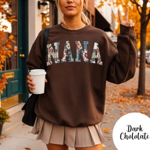 May include: A dark brown sweatshirt with the word "NANA" in a patchwork design of floral and plaid patterns. The sweatshirt is paired with a beige pleated skirt. A white coffee cup is held in the left hand.