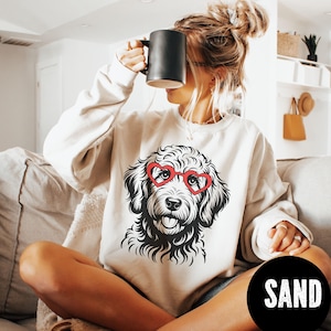 May include: A cream-colored sweatshirt featuring a black and white illustration of a dog wearing red heart-shaped glasses. The word "SAND" is printed in white on a black circle at the bottom right.