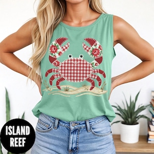 May include: A light green tank top featuring a crab design. The crab has red and white gingham body, red floral claws, and is set against a sandy beach scene. The text "ISLAND REEF" is in a black circle at the bottom.