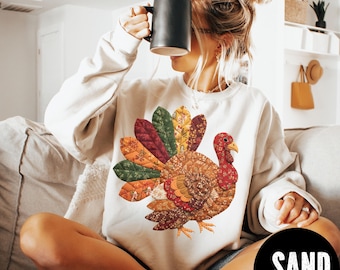 May include: A cream-colored sweatshirt featuring a colorful turkey design. The turkey has a patchwork-style appearance with various autumnal colors. The word "SAND" is printed in white on a black circle at the bottom right.
