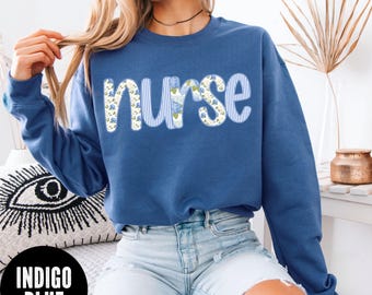 May include: Indigo blue sweatshirt with the word "nurse" in a decorative font. The letters are a combination of blue and white stripes and floral patterns. The sweatshirt is a crew neck style.