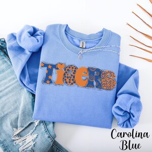 May include: Carolina blue sweatshirt with the word "Tigers" in orange and navy blue patterned letters. The sweatshirt has a crew neck and long sleeves. A silver necklace is draped across the top. The sweatshirt is folded on top of a pair of blue jeans.