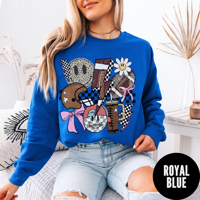 May include: Royal blue sweatshirt with a graphic design featuring a football, helmet, lightning bolt, and smiley faces. The design incorporates checkered patterns, a daisy, and a pink bow. The text "ROYAL BLUE" is in a black circle.